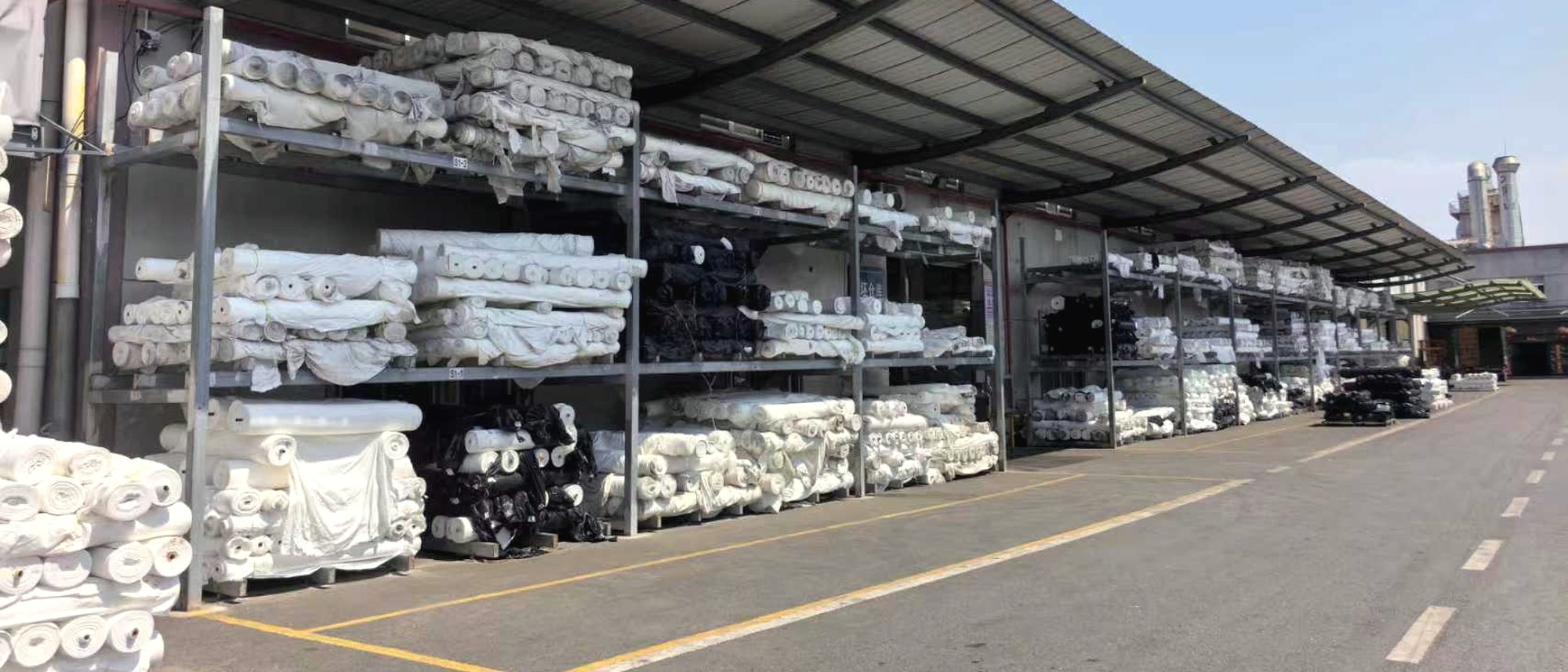 Racks of fabric rolls stacked three high under a covered yard at a partner mill.